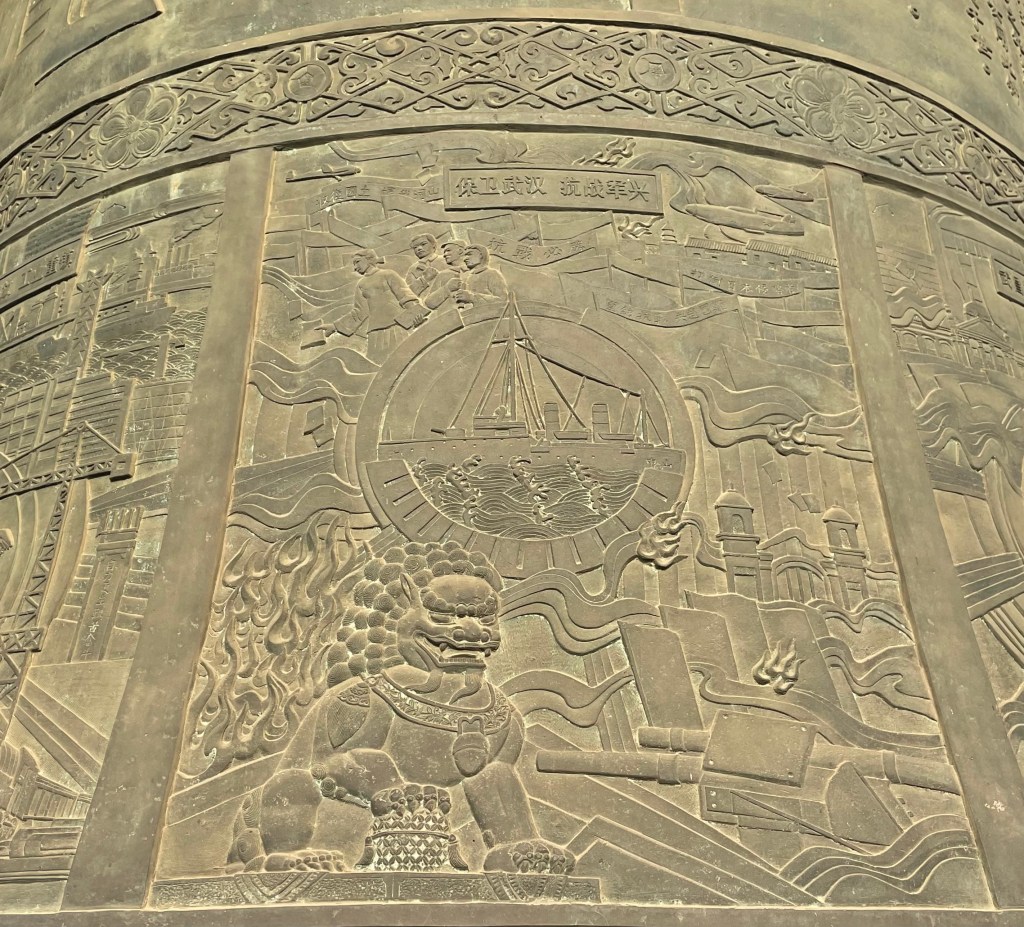 This is a photo of a bell outside Yellow Crane Tower in Wuhan, China. It depicts a Foo Dog and modern industry in Wuhan.