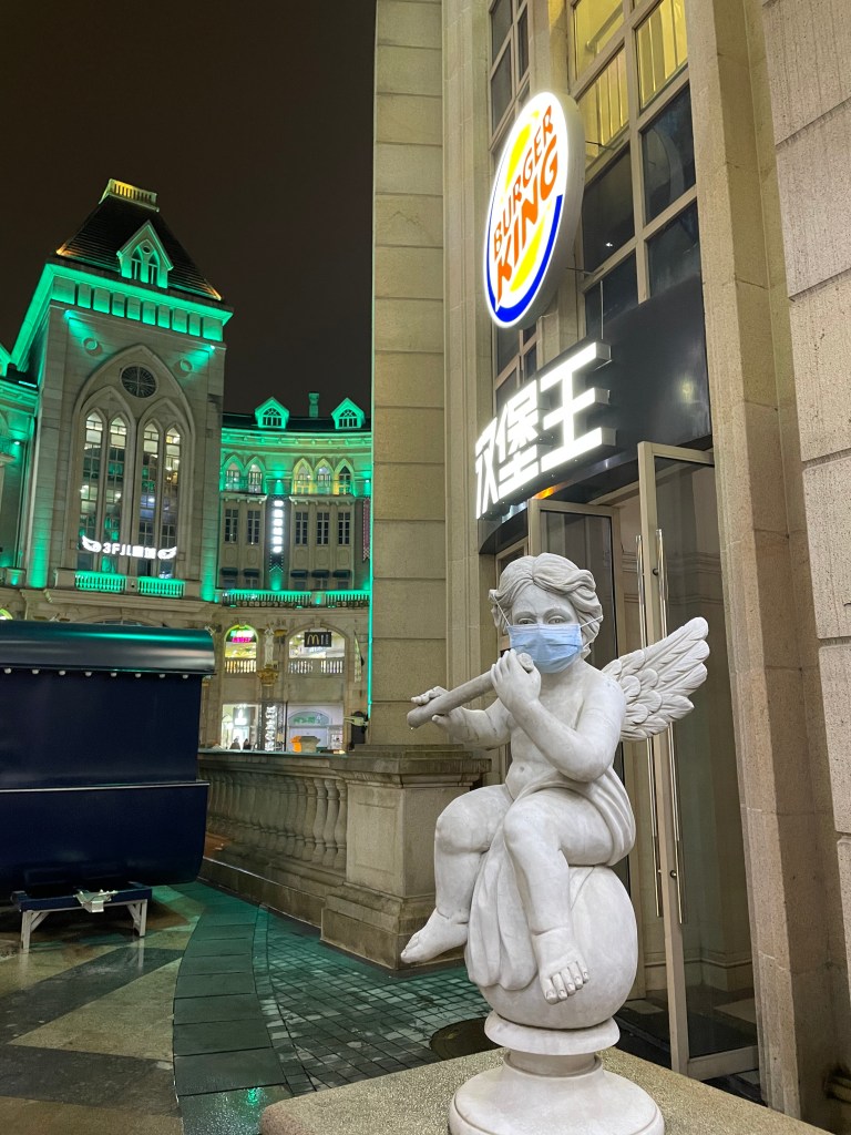 This photo shows an angel statue in front of a Burger King in Wuhan, China in 2021. Someone placed a face mask on the angel as a joke, this photo was taken during the three year long covid-era.