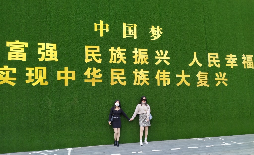 The picture depicts two women posing under a green wall of propaganda in Wuhan, China in 2020.
