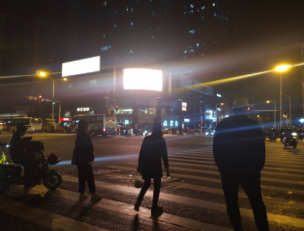 This photo depicts people standing in a crosswalk under neon lights in Wuhan, China (2020).