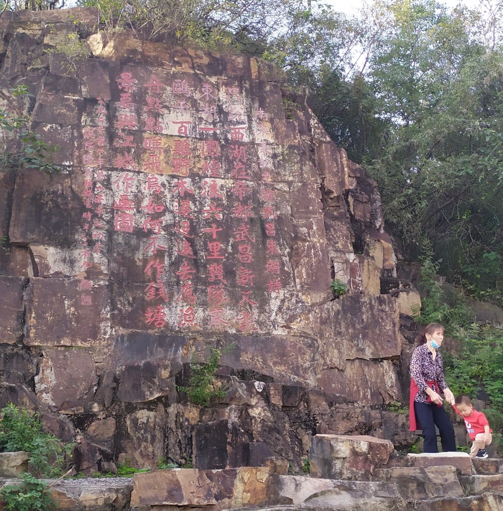 This photo shows a poem written by Mao Zedong carved into a rock facing East Lake in Wuhan, China (2020). A grandmother and grandchild play nearby.