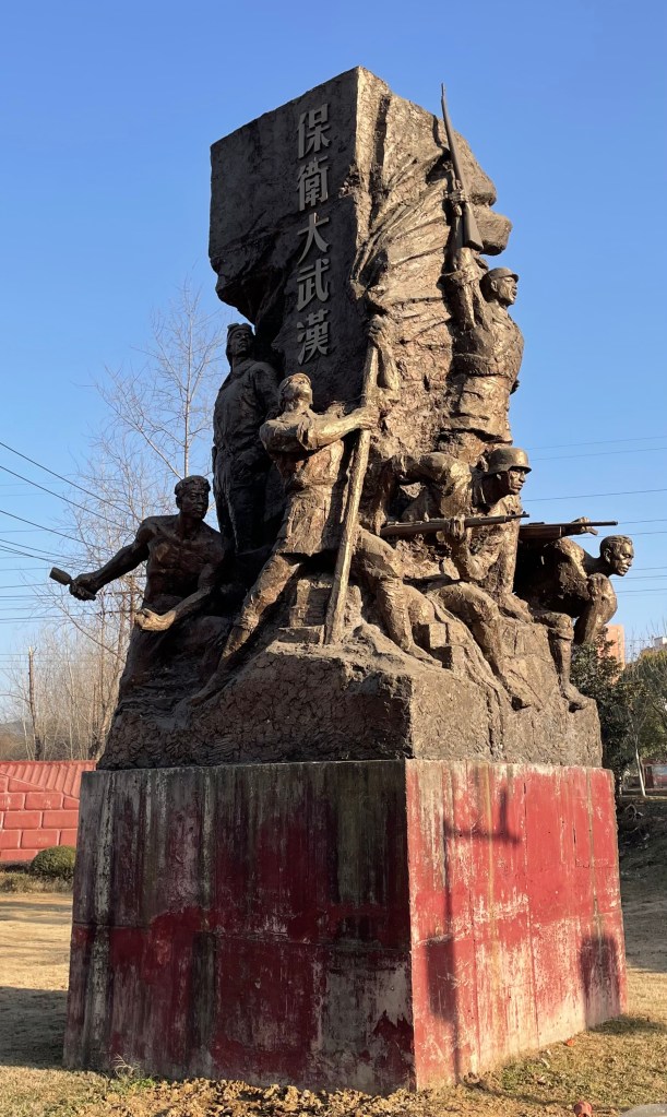 This is a photo of a monument to soldiers in Wuhan, PRC. It says "Defend Big Wuhan". This monument is about 25 feet high and is a fairly typical example of the Soviet-like art that characterizes New China monuments. Taken in January of 2021.