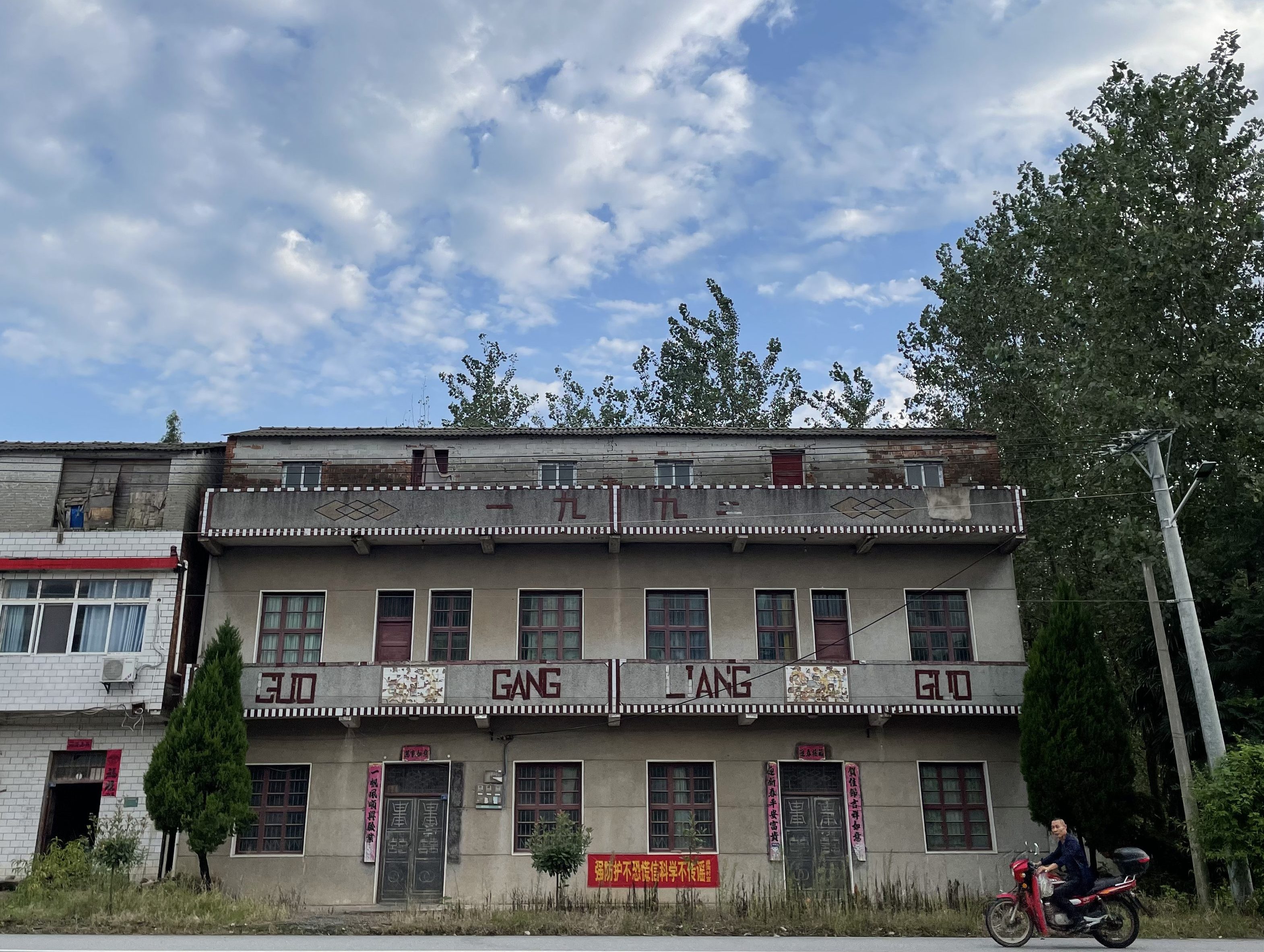 This is a photo of a man riding a motorbike past an older home in Hubei, China in 2021.
