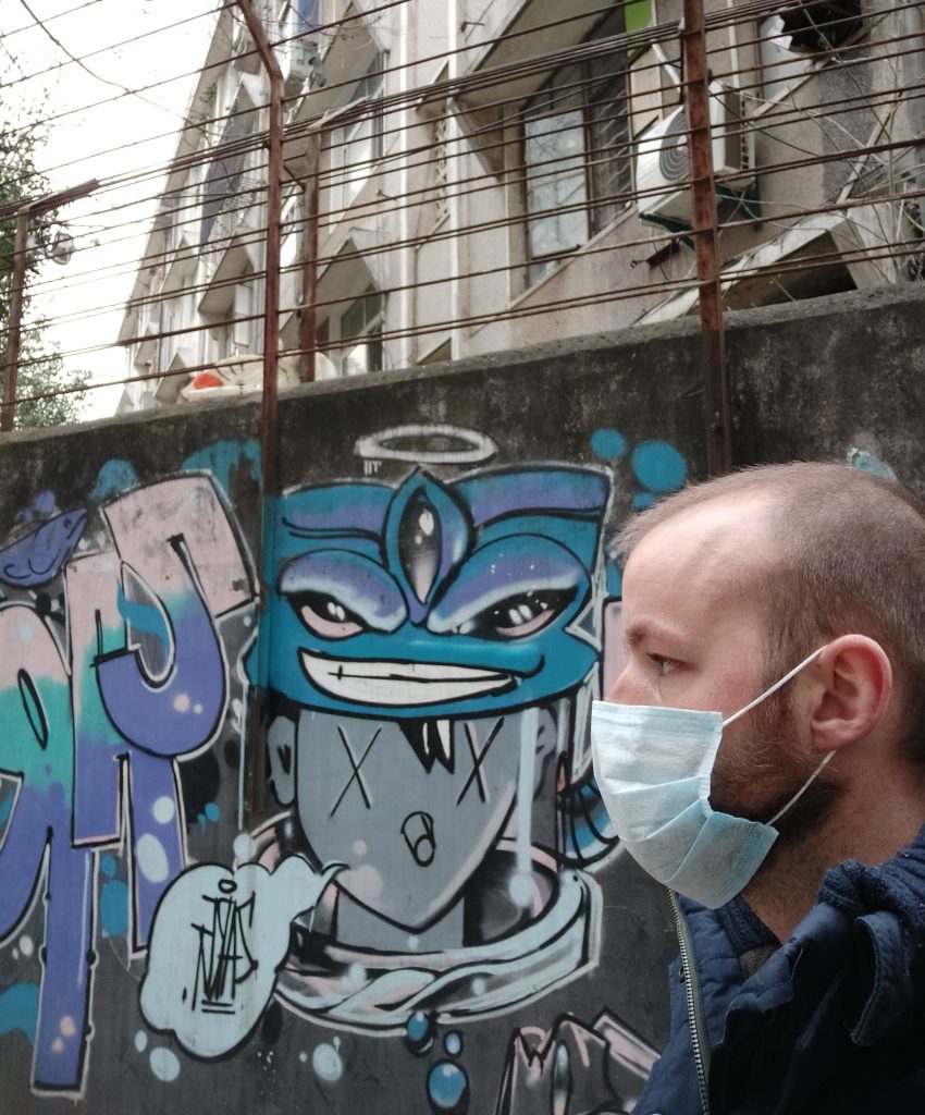 A foreigner in Wuhan in 2020 wearing a mask and standing in front of some graffiti on Wenhua College campus.
